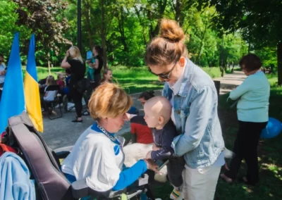 Na środku widać dwie kobiety w parku, są frontem do siebie. Jedna z nich siedzieć na wózku elektrycznym, druga podaje jej na rękach małe dziecko. W tle widać spacerujących ludzi.